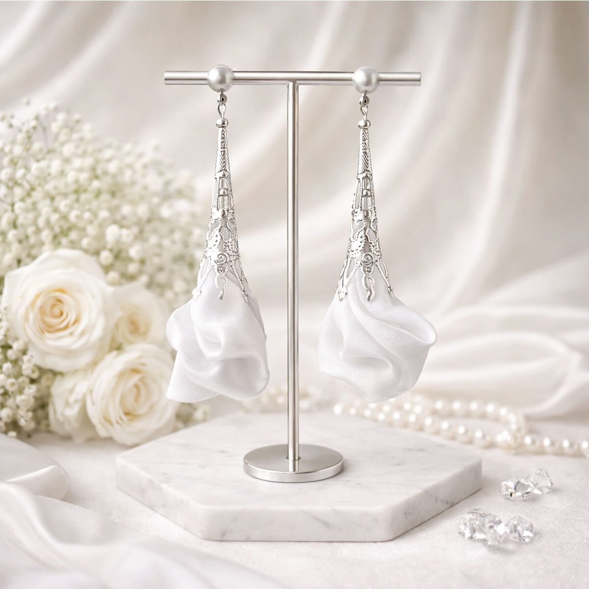 Długie, wiszące kolczyki w kształcie białych kwiatów uformowanych z tkaniny, zwieńczone ażurowym, filigranowym stożkiem w kolorze srebrnym na zapięciu typu sztyft