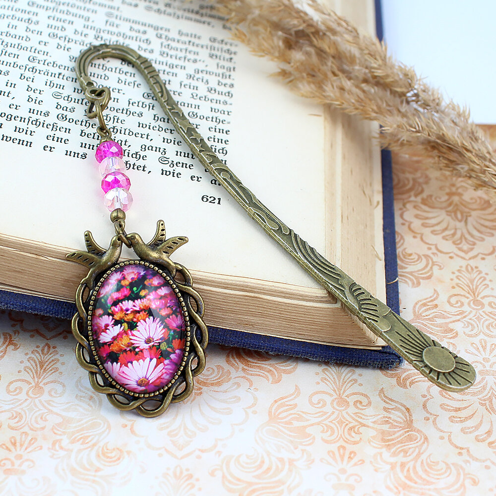 Zakładka do książki z antycznego brązu z różowymi kryształkami i owalnym szklanym kaboszonem z grafiką ptaszków i kwiatów na łące.