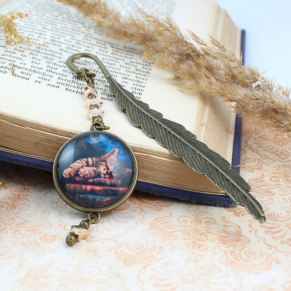 Zakładka do książki z antycznego brązu z zawieszką w kształcie gwiazdek oraz ilustracją śpiącego kota na stosie książek pod szkiełkiem.