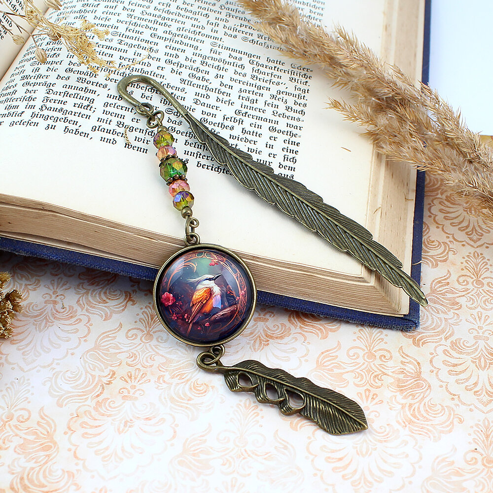 Zakładka do książki z antycznego brązu z szklanym kaboszonem w oprawie zdobionej kryształkami, przedstawiającym ryciny w stylu vintage z sikorą bogatką i zawieszką w kształcie piórka.