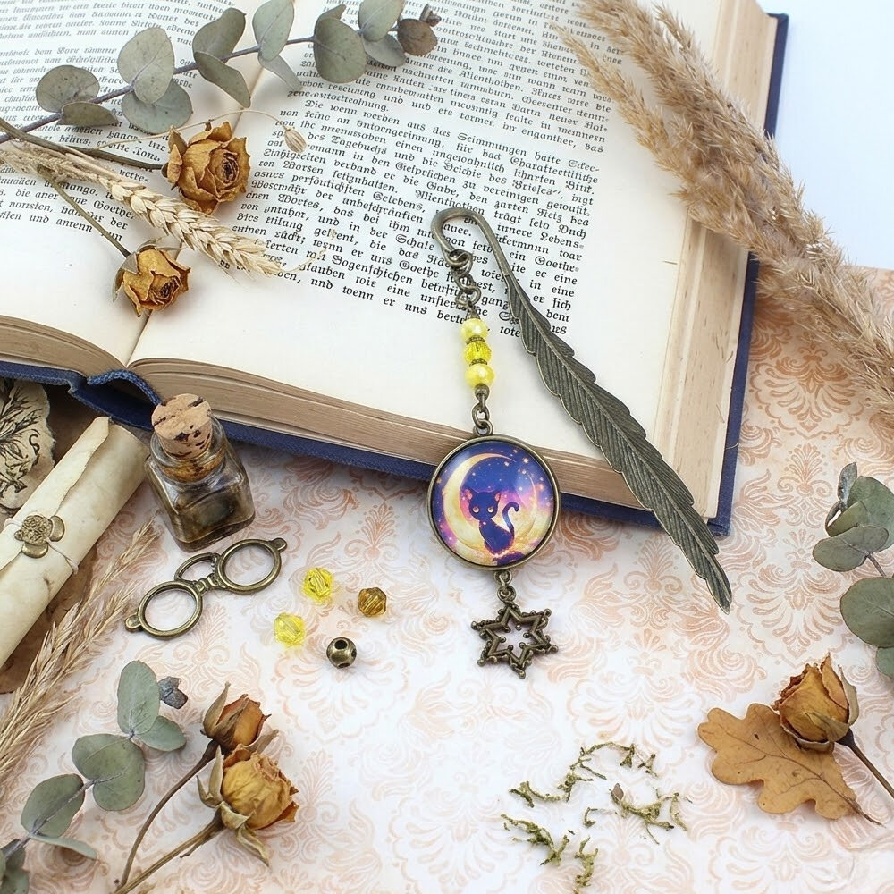 Zakładka do książki w kolorze antycznego brązu w kształcie pióra, z żółtymi kryształkami, zawieszką gwiazdką i wizerunkiem czarnego kota na tle księżyca pod szklanym kaboszonem