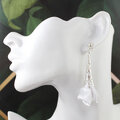 Długie, wiszące kolczyki w kształcie białych kwiatów uformowanych z tkaniny, zwieńczone ażurowym, filigranowym stożkiem w kolorze srebrnym na zapięciu typu sztyft