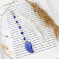 Cienka i lekka metalowa zakładka do książki w kształcie pióra, zdobiona wiszącą zawieszką z szafirowych szklanych kryształków i niebieskiego akrylowego listka, w ozdobnym pudełku. Cienka i lekka metalowa zakładka do książki w kształcie pióra, zdobiona wiszącą zawieszką z szafirowych szklanych kryształków i niebieskiego akrylowego listka, w ozdobnym pudełku.