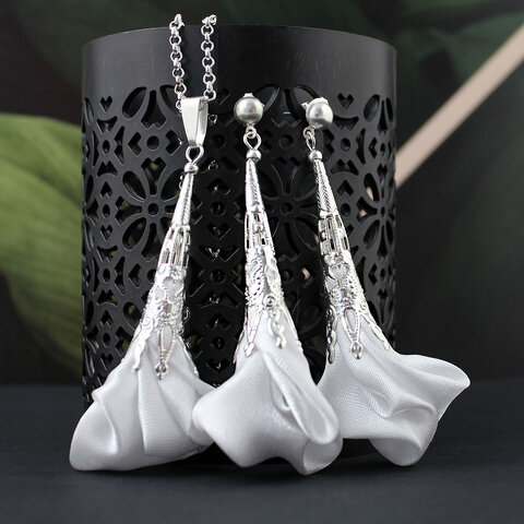 Długie, wiszące kolczyki w kształcie białych kwiatów uformowanych z tkaniny, zwieńczone ażurowym, filigranowym stożkiem w kolorze srebrnym na zapięciu typu sztyft