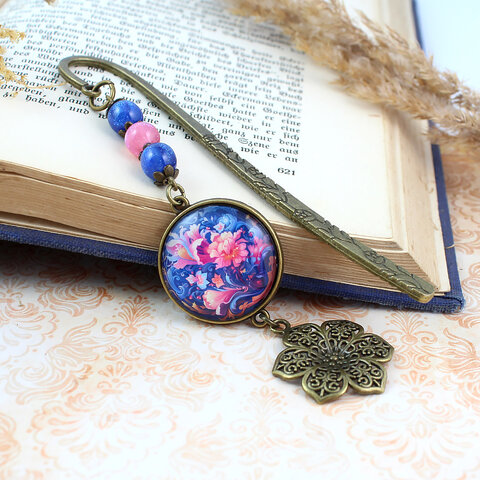 Zakładka do książki z antycznego brązu z niebieskim koralikiem i niebiesko-różowymi, kwitnącymi kwiatami pod szklanym kaboszonem.