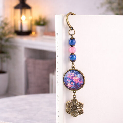 Zakładka do książki z antycznego brązu z niebieskim koralikiem i niebiesko-różowymi, kwitnącymi kwiatami pod szklanym kaboszonem.
